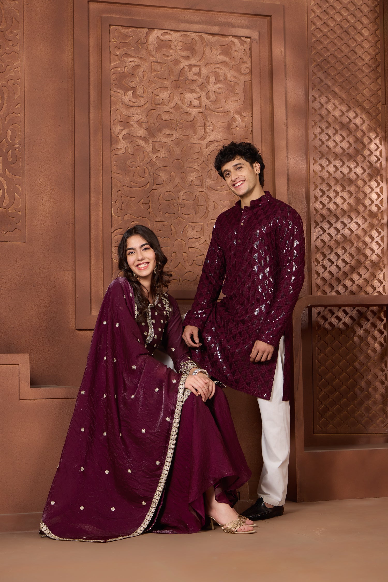 Two people in matching maroon rang collections traditional outfits standing against a decorative brown wall.