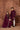Two people in matching maroon rang collections traditional outfits standing against a decorative brown wall.
